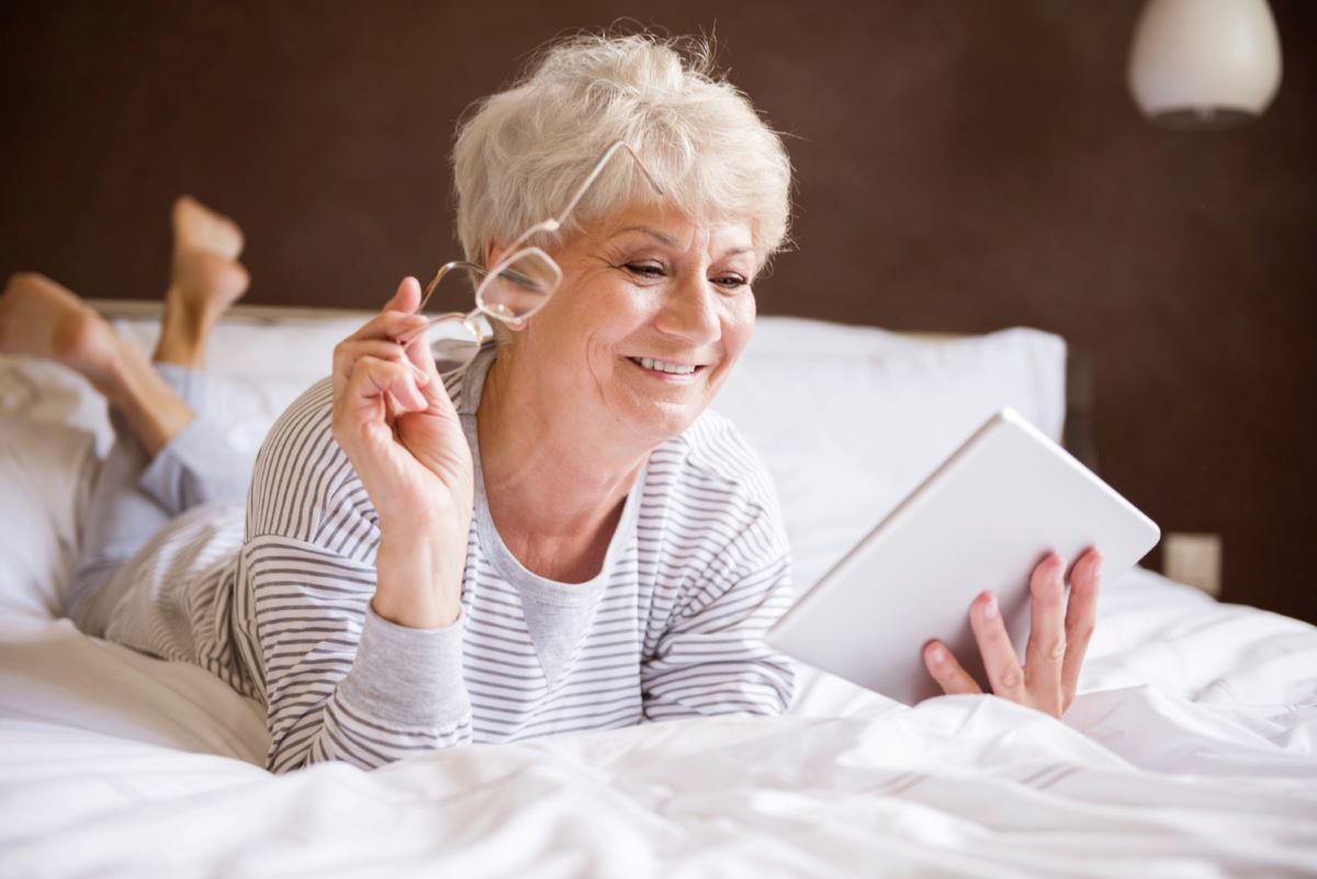 woman on bed holding reading glasses and tablet